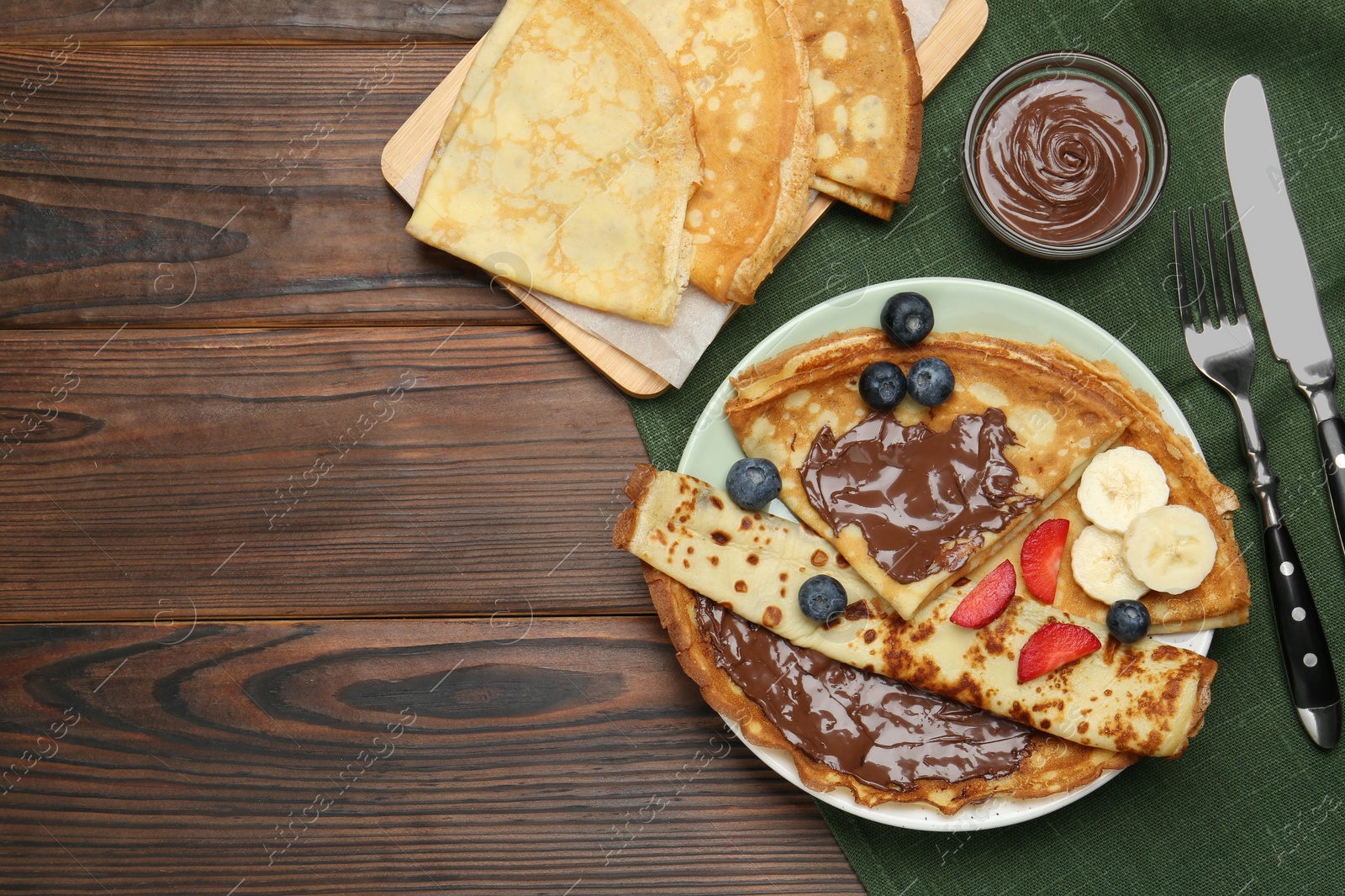 Photo of Tasty crepes with chocolate paste, banana and berries served on wooden table, flat lay. Space for text