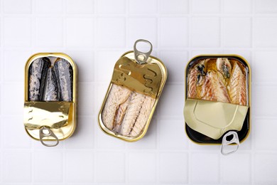 Photo of Open tin cans with mackerel fillets on white tiled table, flat lay