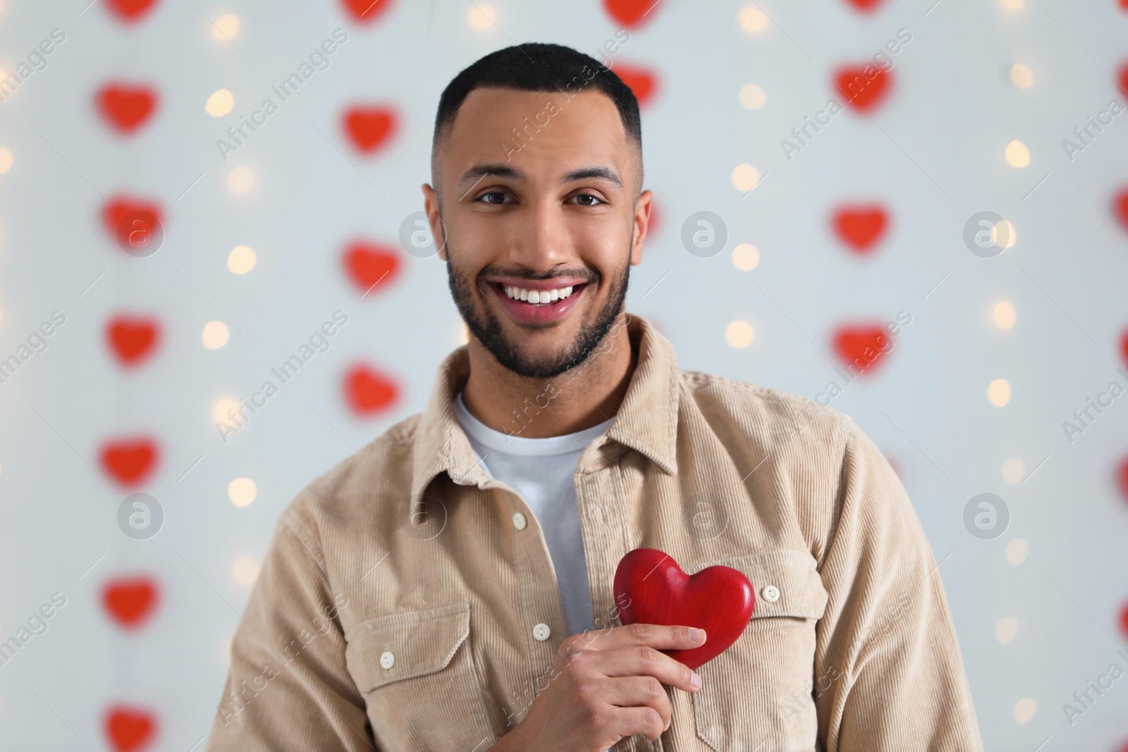 Photo of Handsome young man with red wooden heart indoors, view from camera. Valentine's day celebration in long distance relationship