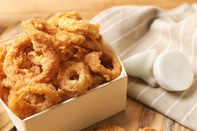 Cardboard box with crunchy fried onion rings on wooden board, closeup