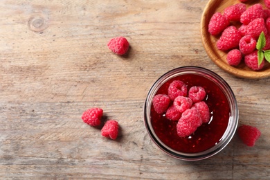 Photo of Glass jar of sweet jam with ripe raspberries on wooden table, flat lay. Space for text