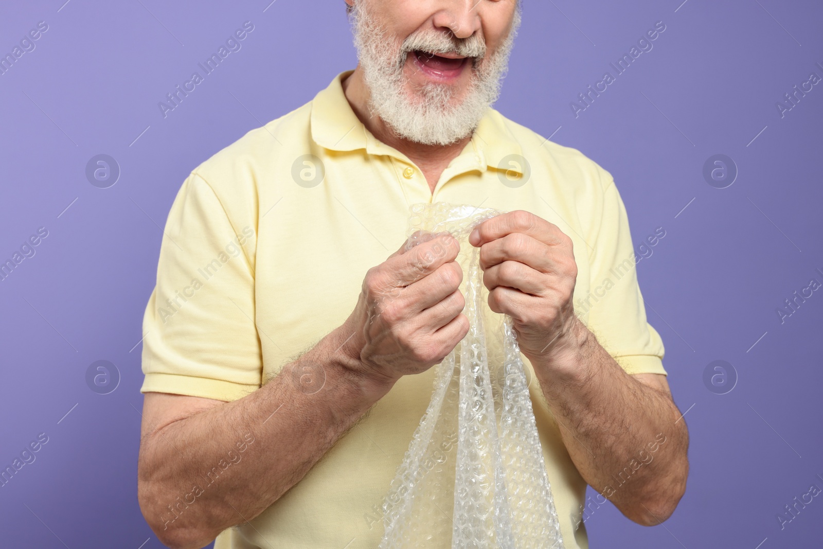 Photo of Senior man popping bubble wrap on light purple background, closeup. Stress relief