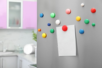 Photo of Empty sheet of paper with colorful magnets on refrigerator door in kitchen. Space for text