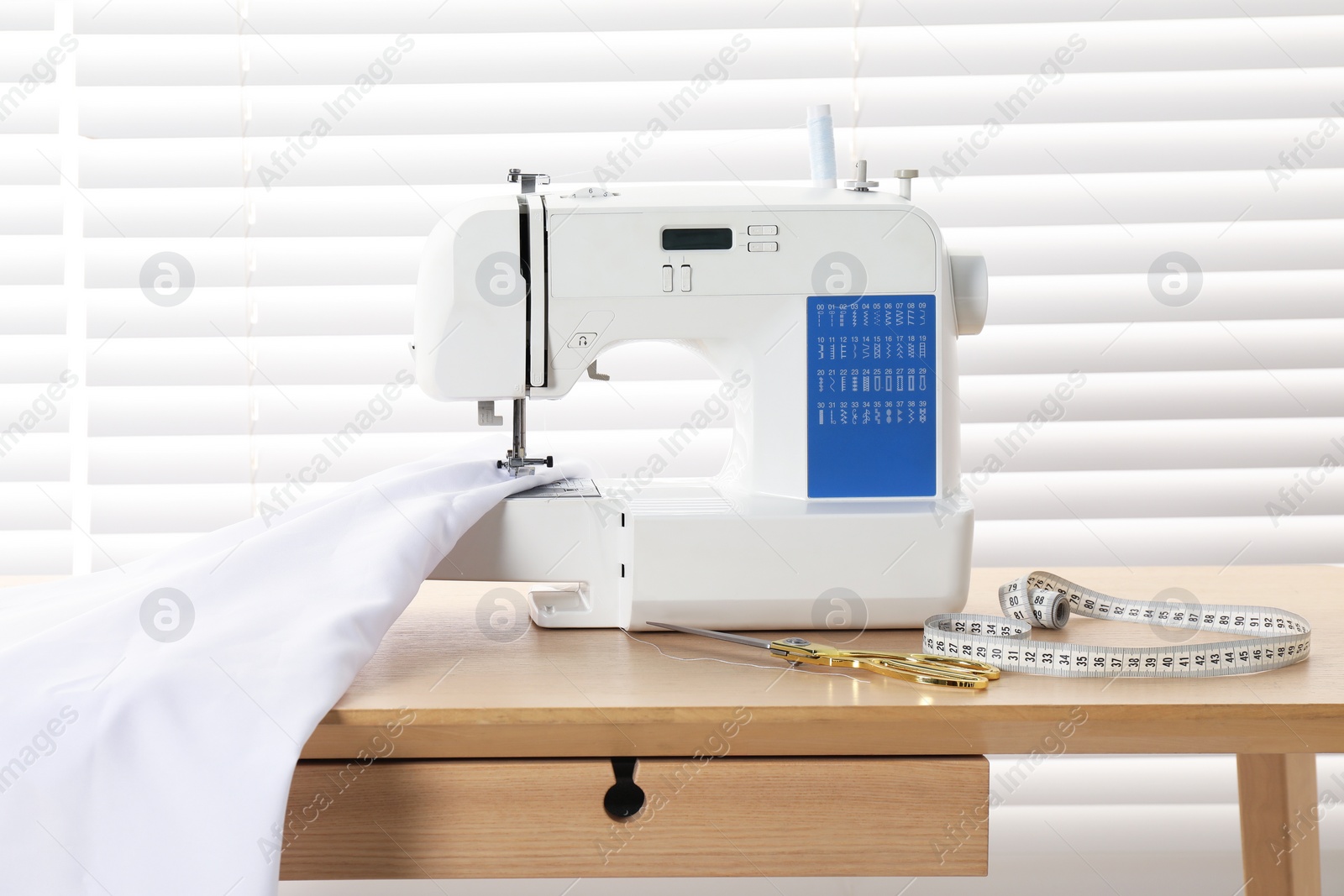 Photo of White sewing machine, scissors, measuring tape and fabric on wooden table indoors