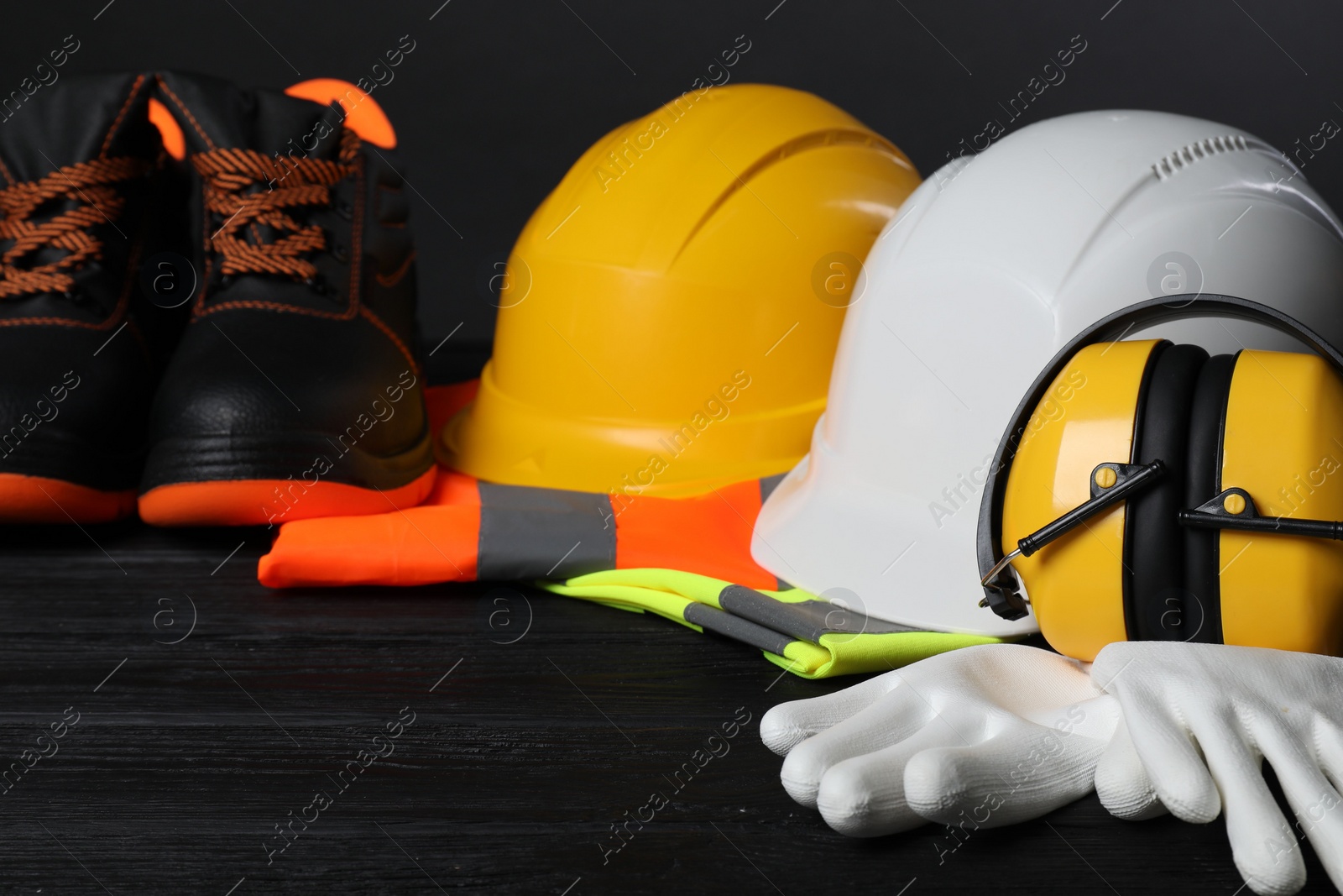 Photo of Pair of working boots, hard hats, protective gloves, earmuffs and reflective vests on black wooden surface, closeup