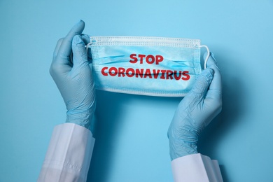 Doctor holding medical mask with text Stop Coronavirus on light blue background, closeup. Protective measures during pandemic