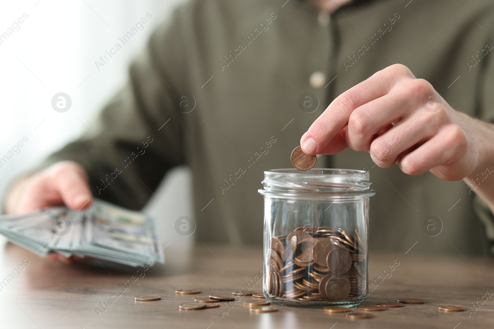 Photo of Financial savings. Man putting coin into glass jar at wooden table, closeup