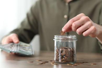 Financial savings. Man putting coin into glass jar at wooden table, closeup