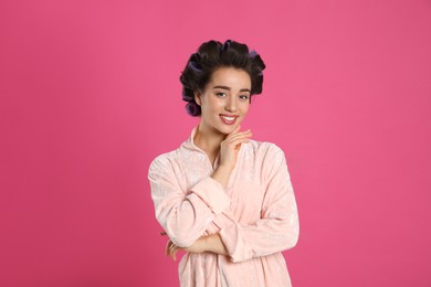 Photo of Happy young woman in bathrobe with hair curlers on pink background