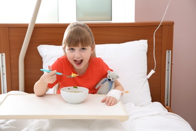 Photo of Little child with intravenous drip eating soup in hospital bed