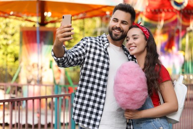 Photo of Happy young man and his girlfriend with cotton candy taking selfie at funfair