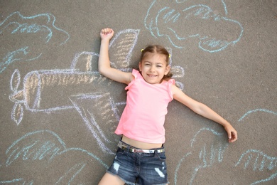 Photo of Little child lying near chalk drawing of airplane on asphalt, top view