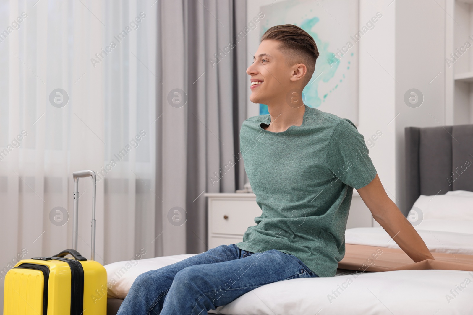 Photo of Smiling guest with suitcase relaxing on bed in stylish hotel room