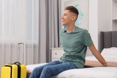 Photo of Smiling guest with suitcase relaxing on bed in stylish hotel room