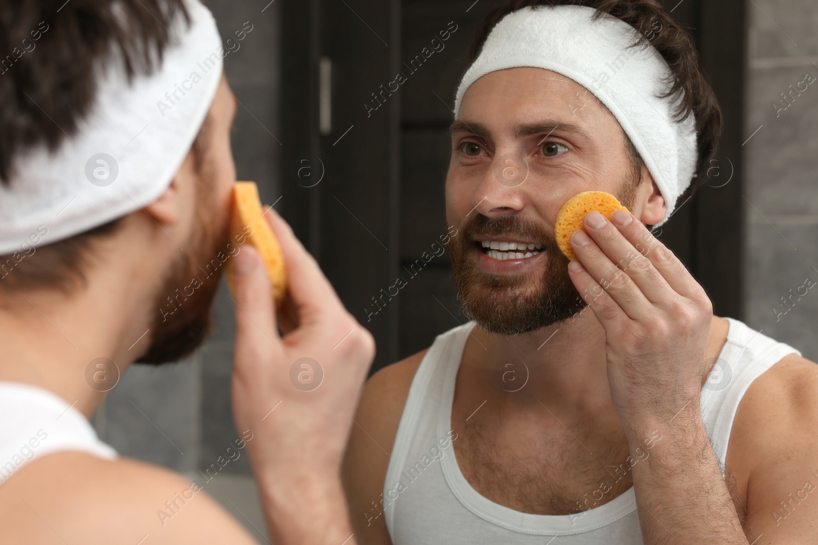 Photo of Man with headband washing his face using sponge near mirror in bathroom