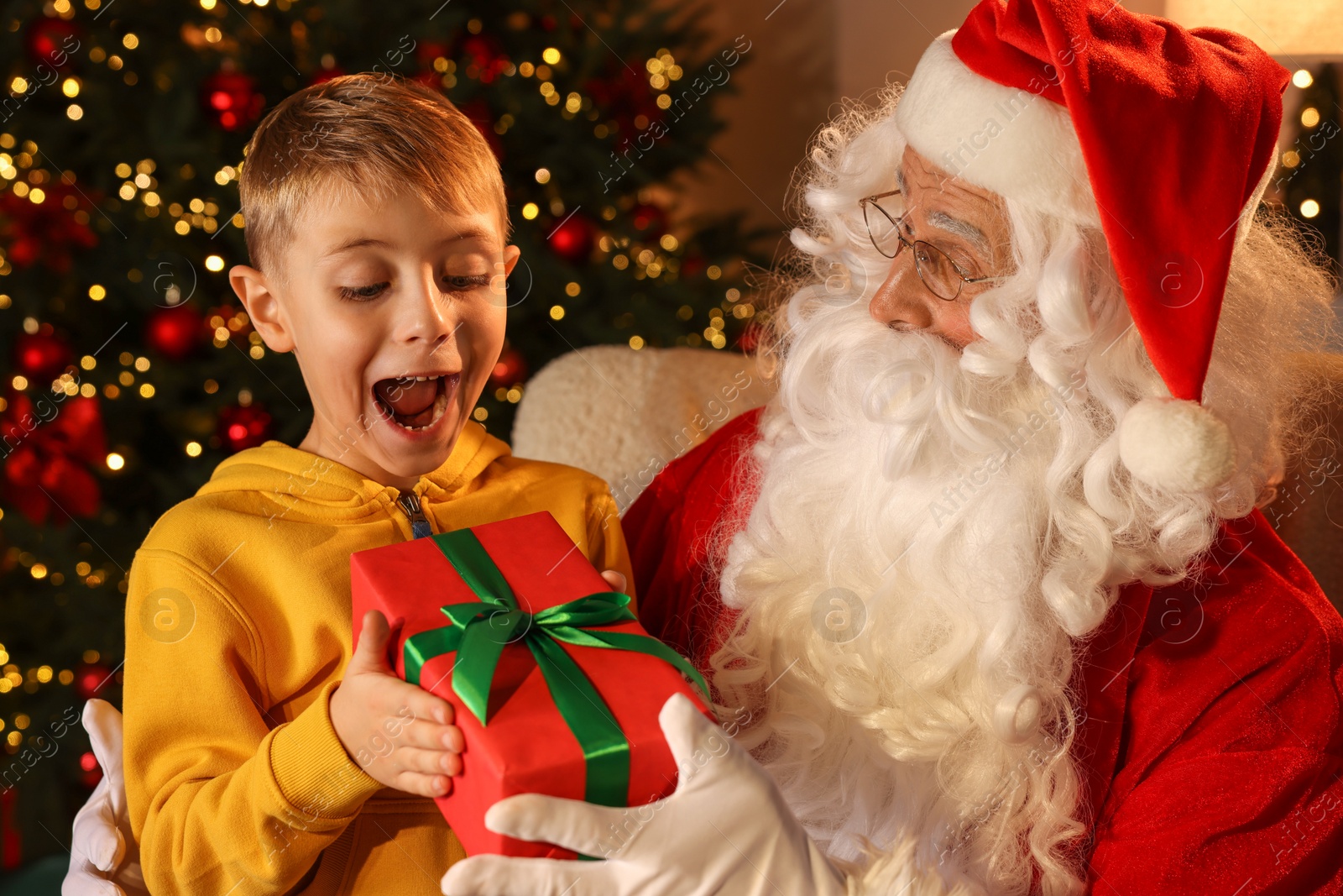 Photo of Surprised little boy taking gift from Santa Claus in room with Christmas tree