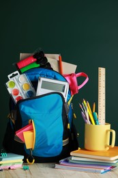 Backpack with different school stationery on white wooden table near chalkboard