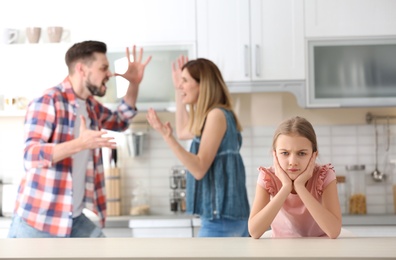 Photo of Little unhappy girl sitting at table while parents arguing on kitchen