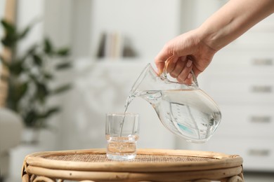 Woman pouring fresh water from jug into glass at wicker surface against blurred background, closeup