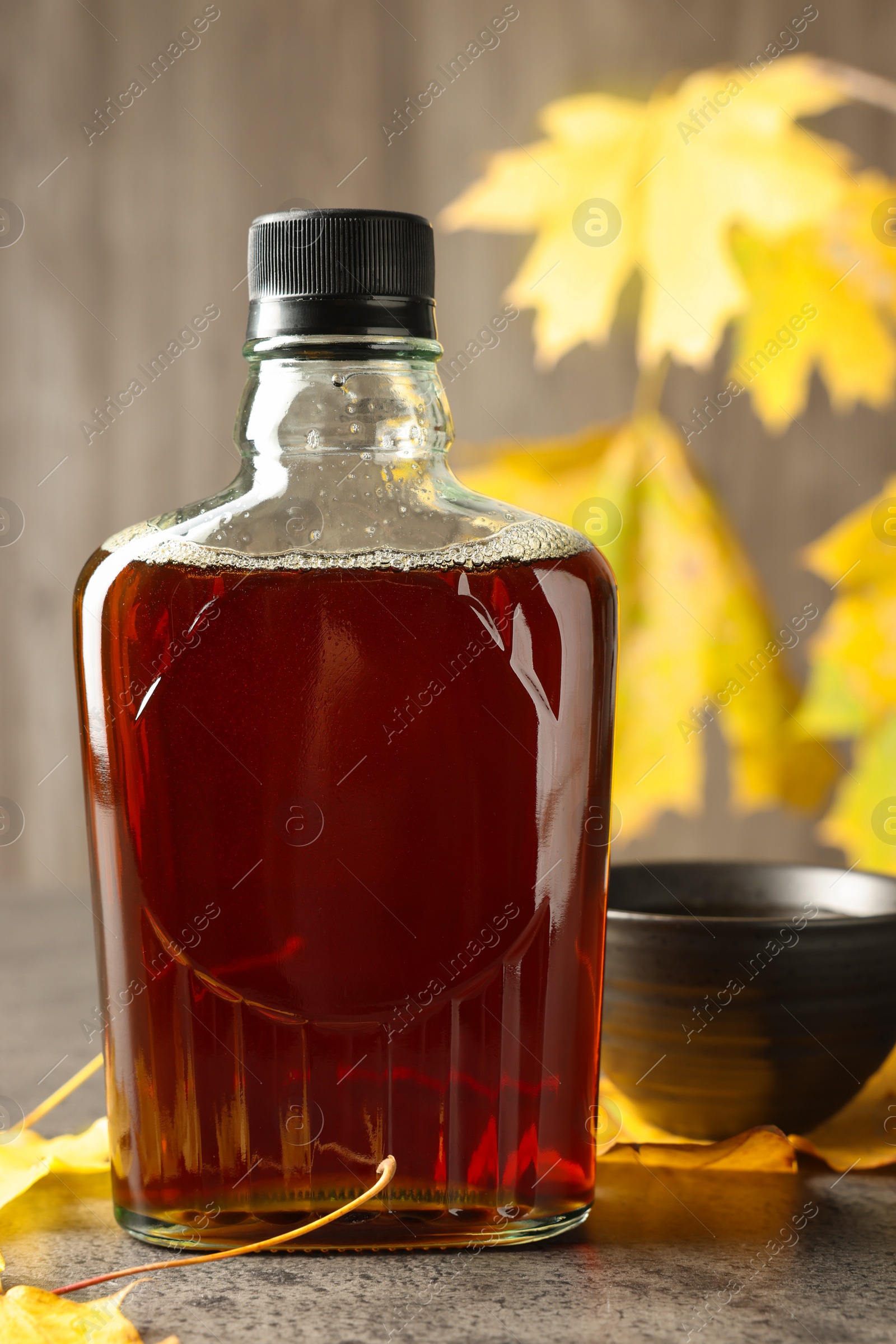 Photo of Bottle of tasty maple syrup, bowl and dry leaves on light grey table