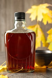 Bottle of tasty maple syrup, bowl and dry leaves on light grey table