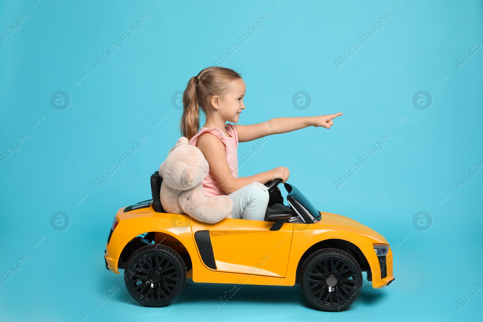 Photo of Cute little girl with toy bear driving children's car on light blue background