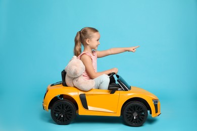 Cute little girl with toy bear driving children's car on light blue background