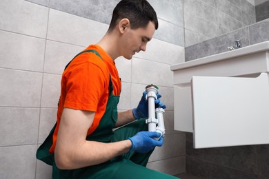 Young plumber wearing protective gloves repairing sink in bathroom