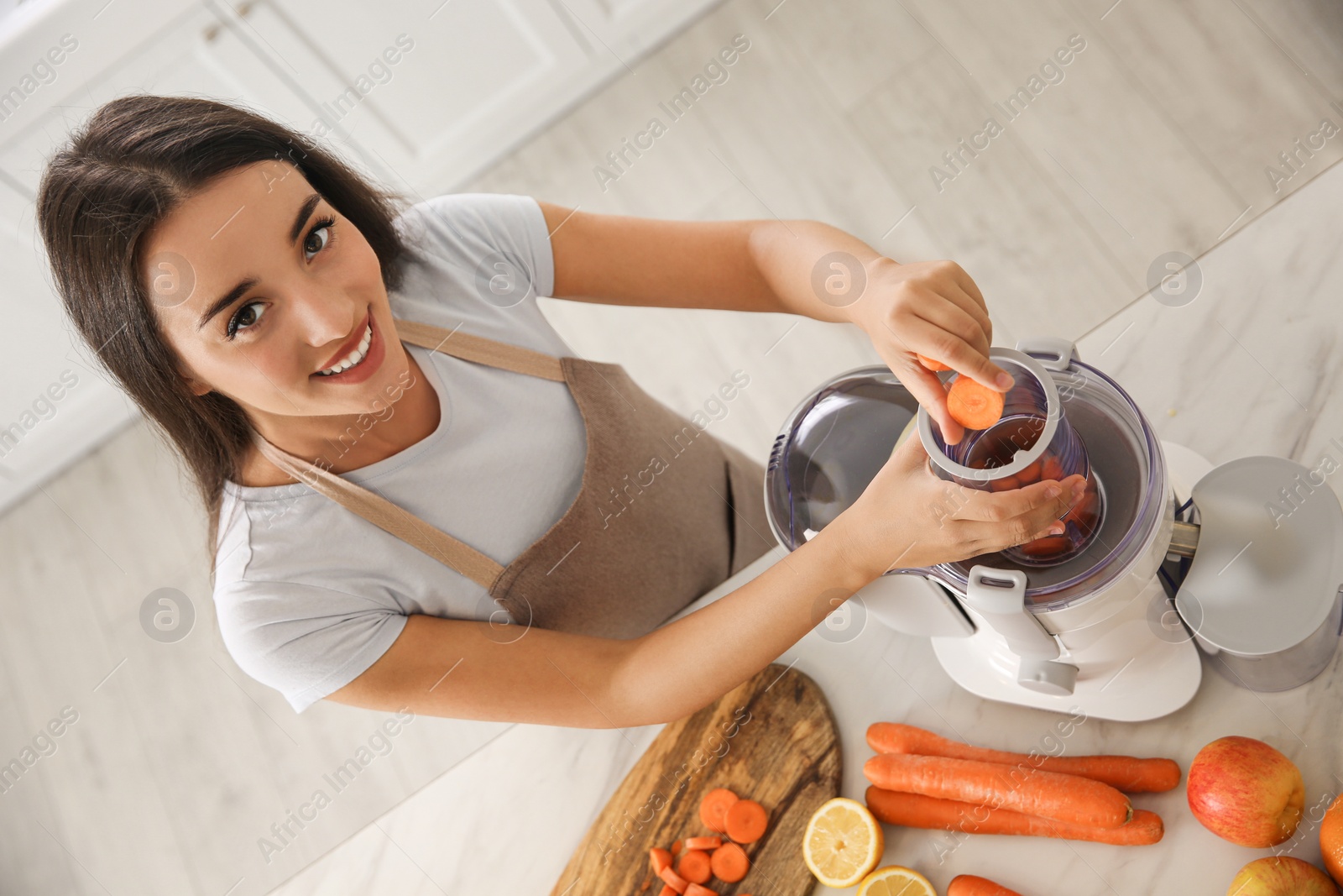 Photo of Young woman putting fresh slices of carrot into juicer at table in kitchen, above view