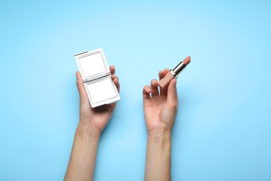 Photo of Woman holding pocket mirror and lipstick on light blue background, top view