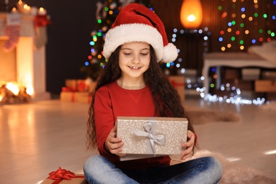 Photo of Cute little child in Santa hat with Christmas gift box at home