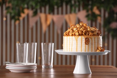 Photo of Caramel drip cake decorated with popcorn and pretzels near tableware on wooden table