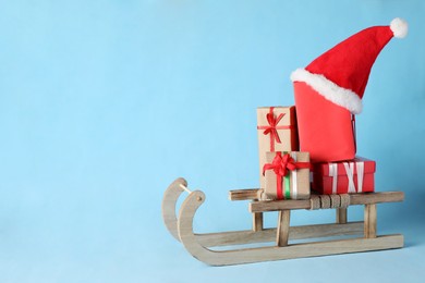 Photo of Wooden sleigh with Christmas gift boxes and Santa hat on light blue background, space for text