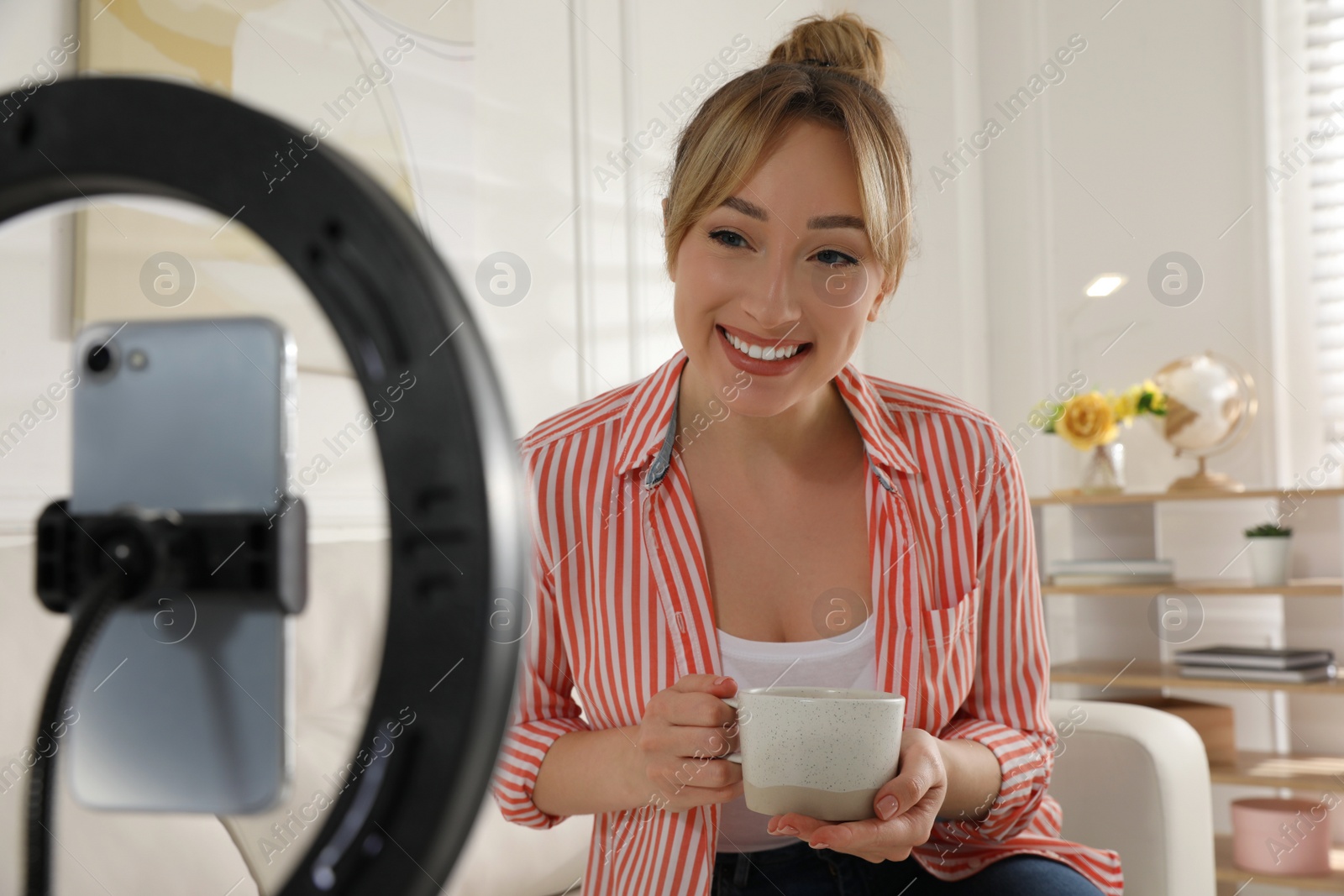 Photo of Blogger with cup of tea recording video in living room at home. Using ring lamp and smartphone