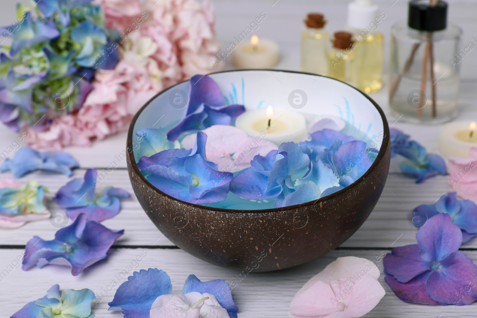 Photo of Spa composition. Aromatic water with burning candle in bowl and flowers on white wooden table, closeup