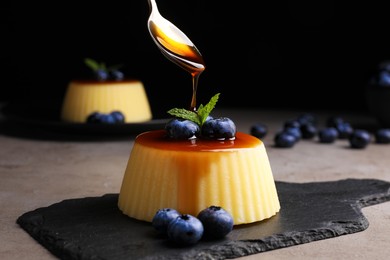 Pouring caramel onto delicious pudding with blueberries and mint on grey table against black background, closeup