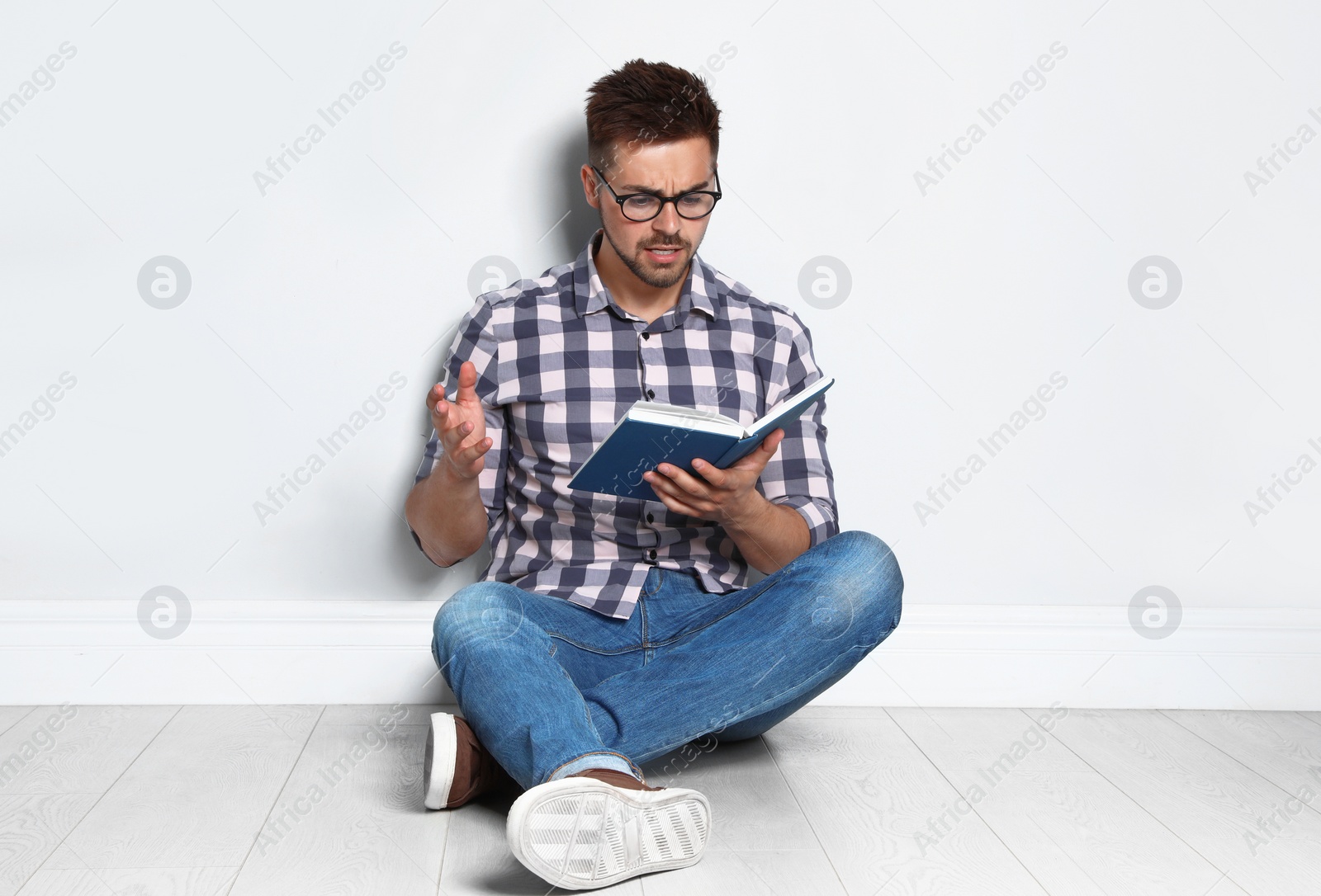 Photo of Handsome young man reading book on wooden floor near light wall