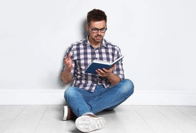 Handsome young man reading book on wooden floor near light wall