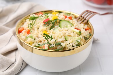 Cooked bulgur with vegetables in bowl on white tiled table, closeup