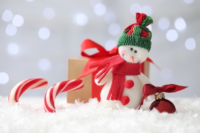 Photo of Snowman toy, candies and Christmas ball on snow against blurred festive lights