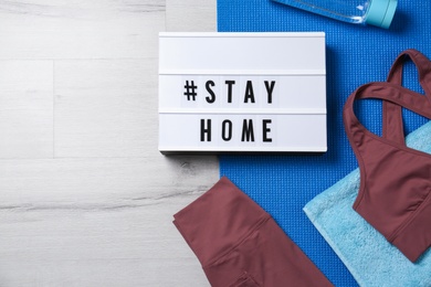 Photo of Stylish sportswear, yoga mat and light box with hashtag Stay at Home on white wooden background, flat lay. Self isolation during COVID‑19 pandemic