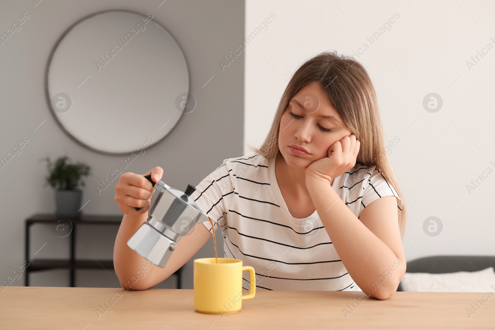 Photo of Sleepy young woman pouring coffee into cup at wooden table indoors