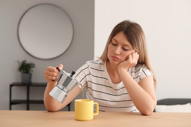 Sleepy young woman pouring coffee into cup at wooden table indoors