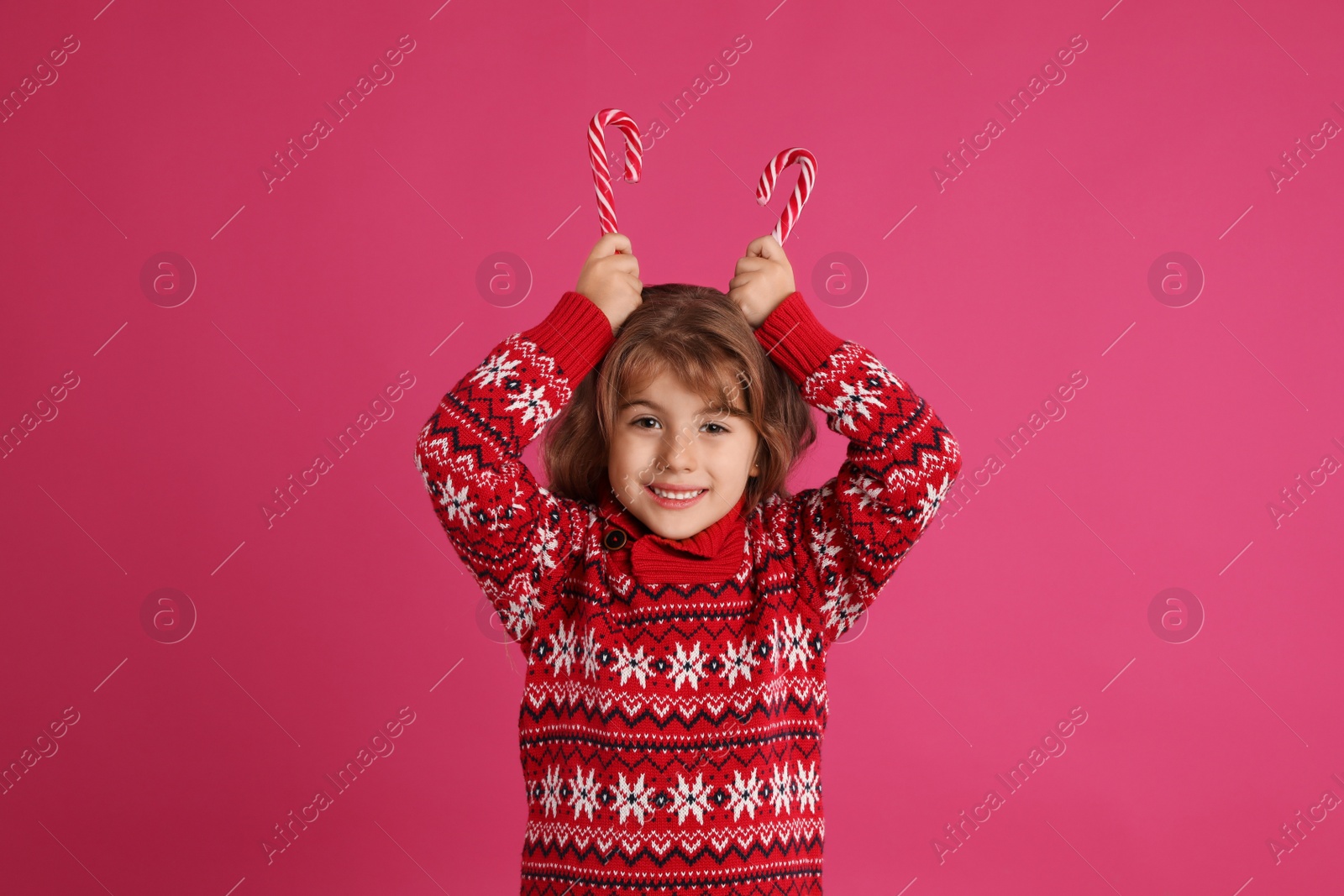 Photo of Cute little girl in Christmas sweater holding sweet candy canes near head against pink background