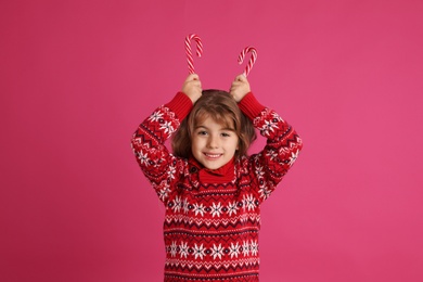 Photo of Cute little girl in Christmas sweater holding sweet candy canes near head against pink background