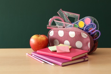 Photo of Different school stationery and apple on wooden table near green chalkboard. Back to school