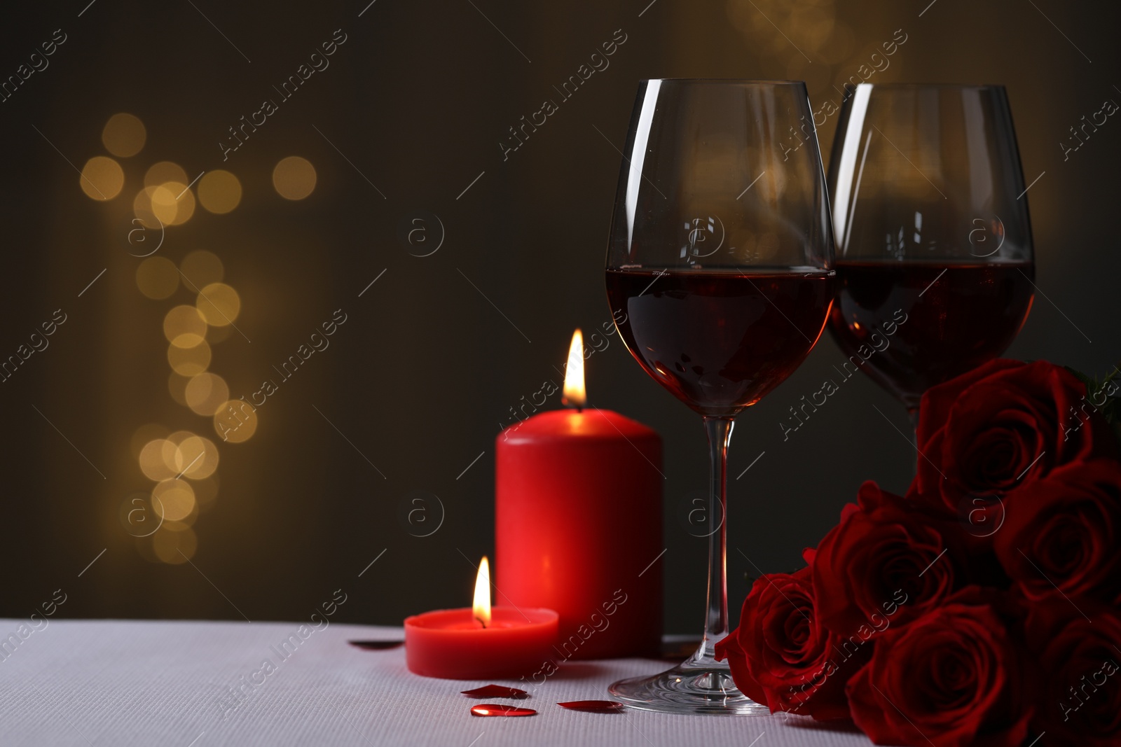 Photo of Glasses of red wine, burning candles and rose flowers on grey table against blurred lights, space for text. Romantic atmosphere