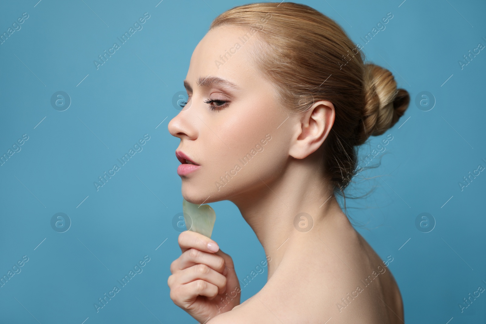 Photo of Beautiful young woman doing facial massage with gua sha tool on blue background, closeup