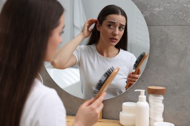 Emotional woman with brush suffering from dandruff near mirror at home.