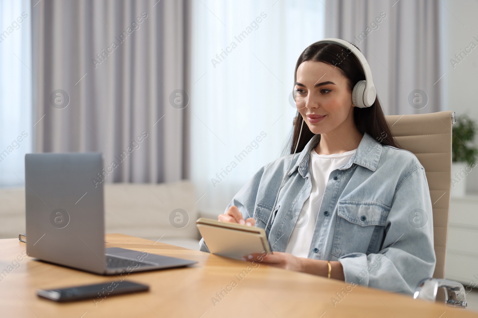 Photo of Young woman in headphones watching webinar at table in room